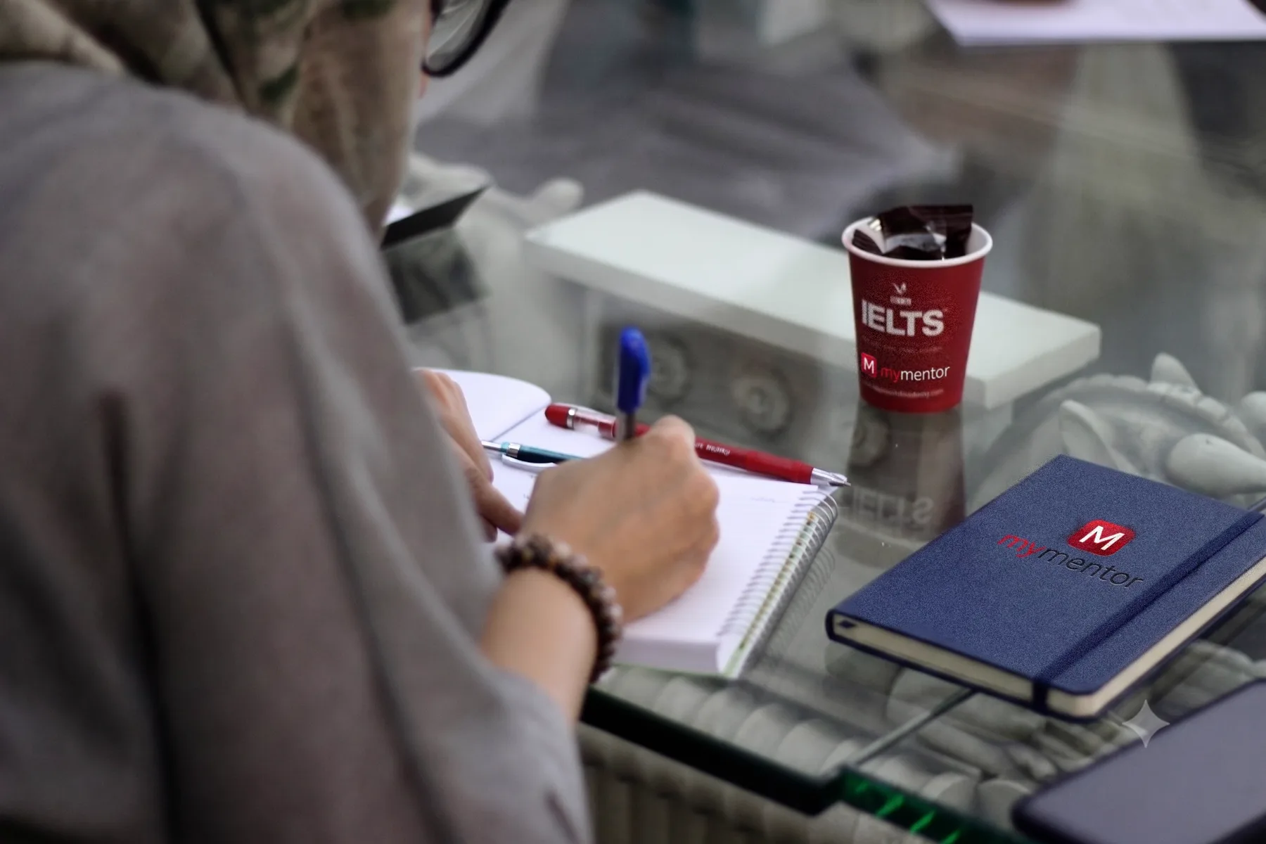 A student taking notes at a desk with a MyMentor notebook and IELTS cup